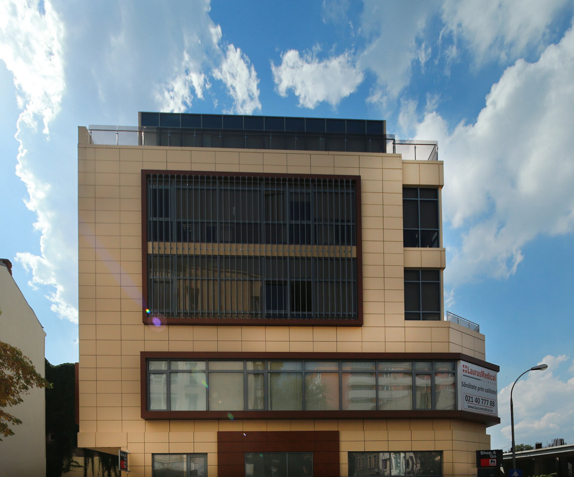 Main facade of the Mihai Eminescu 100 building with ceramic cladding in warm tones, vertical sunshades on the upper floor, a wood-profile finish on horizontal panels and the „Laurus Medical Clinic” sign on the ground floor — a B+G+4F building, PPAE project, initial clinic phase